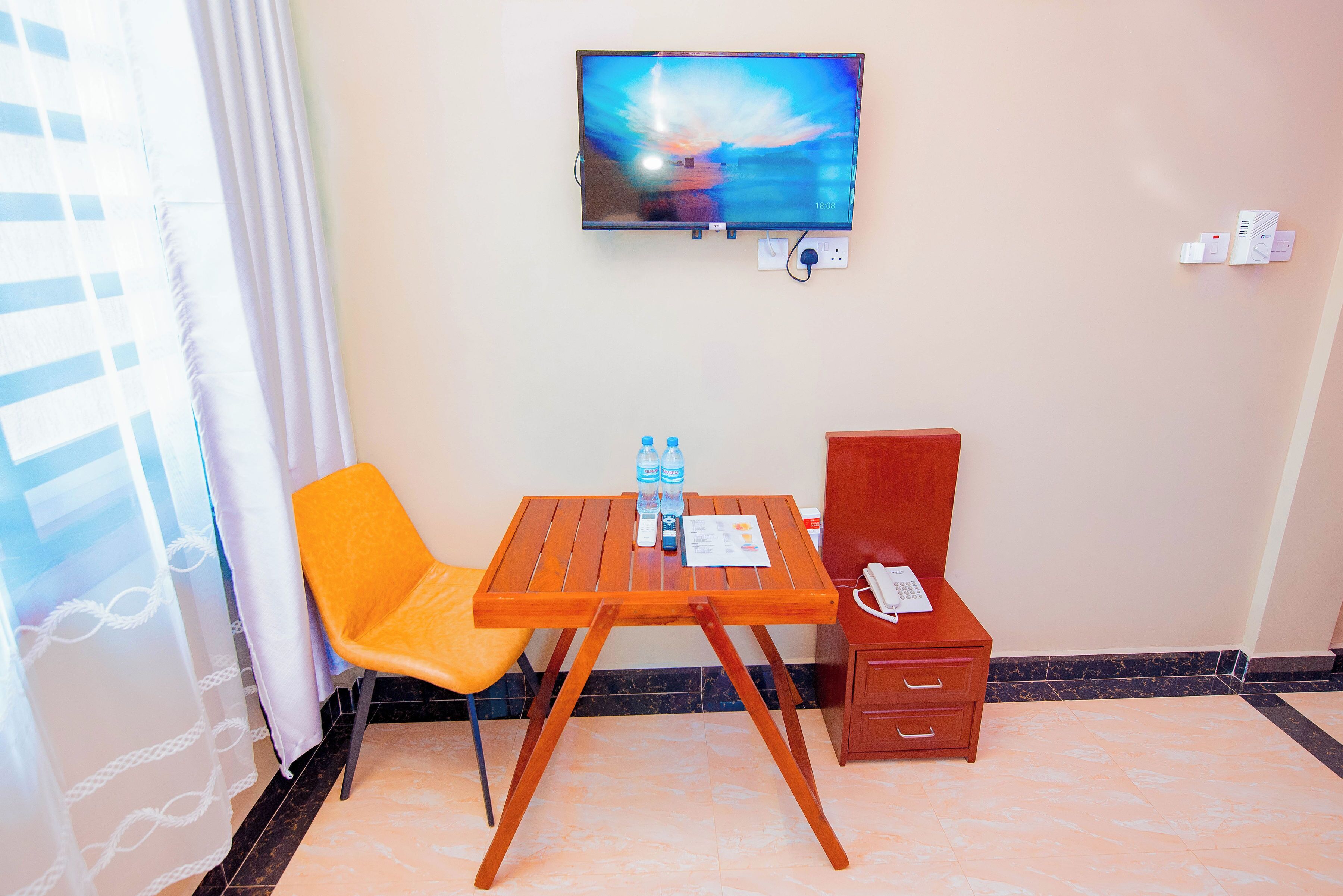 Una habitación de hotel con televisión montada en la pared, una mesa de madera con dos sillas, una cómoda pequeña y un ventanal con cortinas.