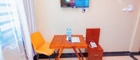 Una habitación de hotel con televisión montada en la pared, una mesa de madera con dos sillas, una cómoda pequeña y un ventanal con cortinas.
