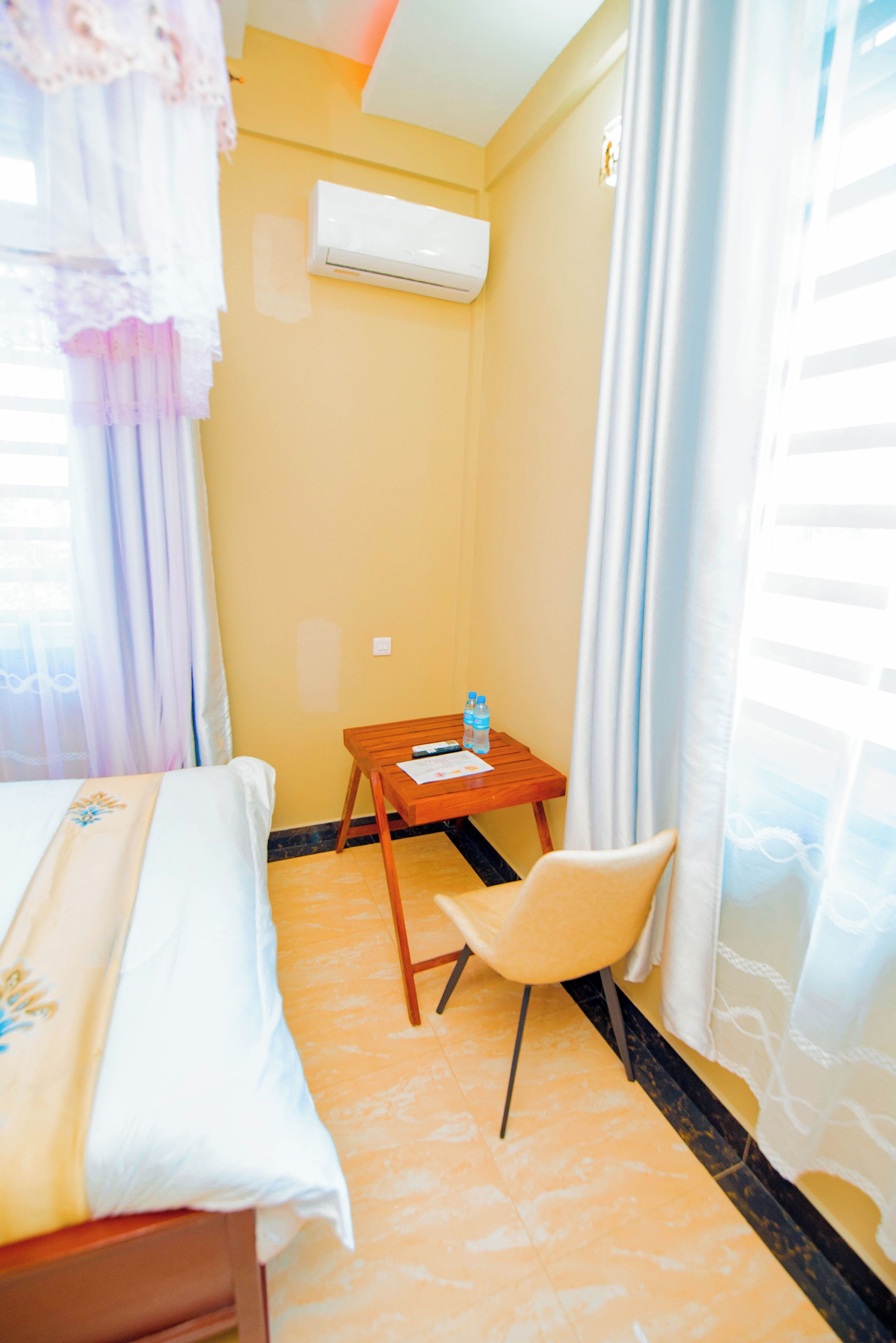 A hotel room with a bed, a small table with a bottle, a chair, and an air conditioning unit on the wall.