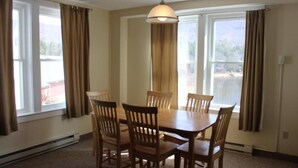 A dining room with a wooden table and chairs, large windows with curtains, and a hanging light fixture.