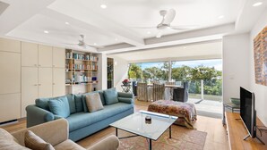 En rummelig stue med blå sofa, sofabord i glas og fjernsyn. Der er et bogreol fyldt med bøger og en balkon med udendørs møbler.