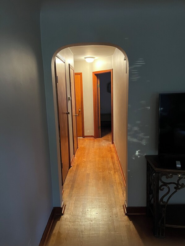 A narrow hallway with wooden flooring and a curved ceiling, leading to a room with a television and a small table.