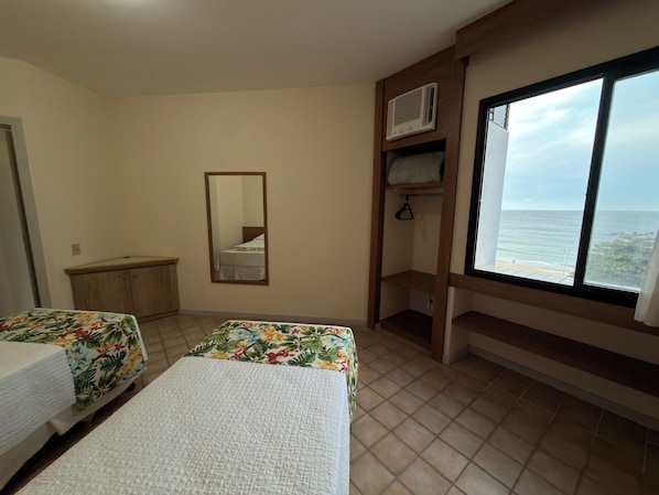 A hotel room with two beds, a mirror, a wardrobe, a shelf with a hanging item, and a window with a view of the sea.