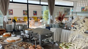 A buffet with various pastries and dishes, a seating area with tables and chairs, and large windows with curtains. - Ingleses Praia Hotel (Florianópolis)