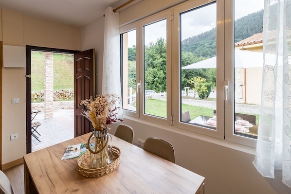 A dining area with a wooden table, chairs, a vase with dried flowers, and large windows offering a view of a garden and mountains.