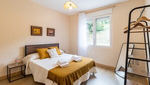 Ein Schlafzimmer mit einem Bett, Nachttischen, einer Pendelleuchte, einem Fenster mit Vorhängen und einem Metallregal.