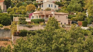 A hillside villa with a terracotta roof, surrounded by lush greenery and other residential buildings.