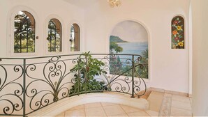 A grand staircase with ornate metal railings, a view of the sea through a mural, and a chandelier.