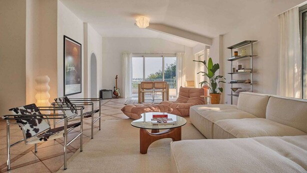 A modern living room with a sofa, a glass coffee table, and a metal bench with a leather seat. There is a large window with curtains, a plant, and a bookshelf.