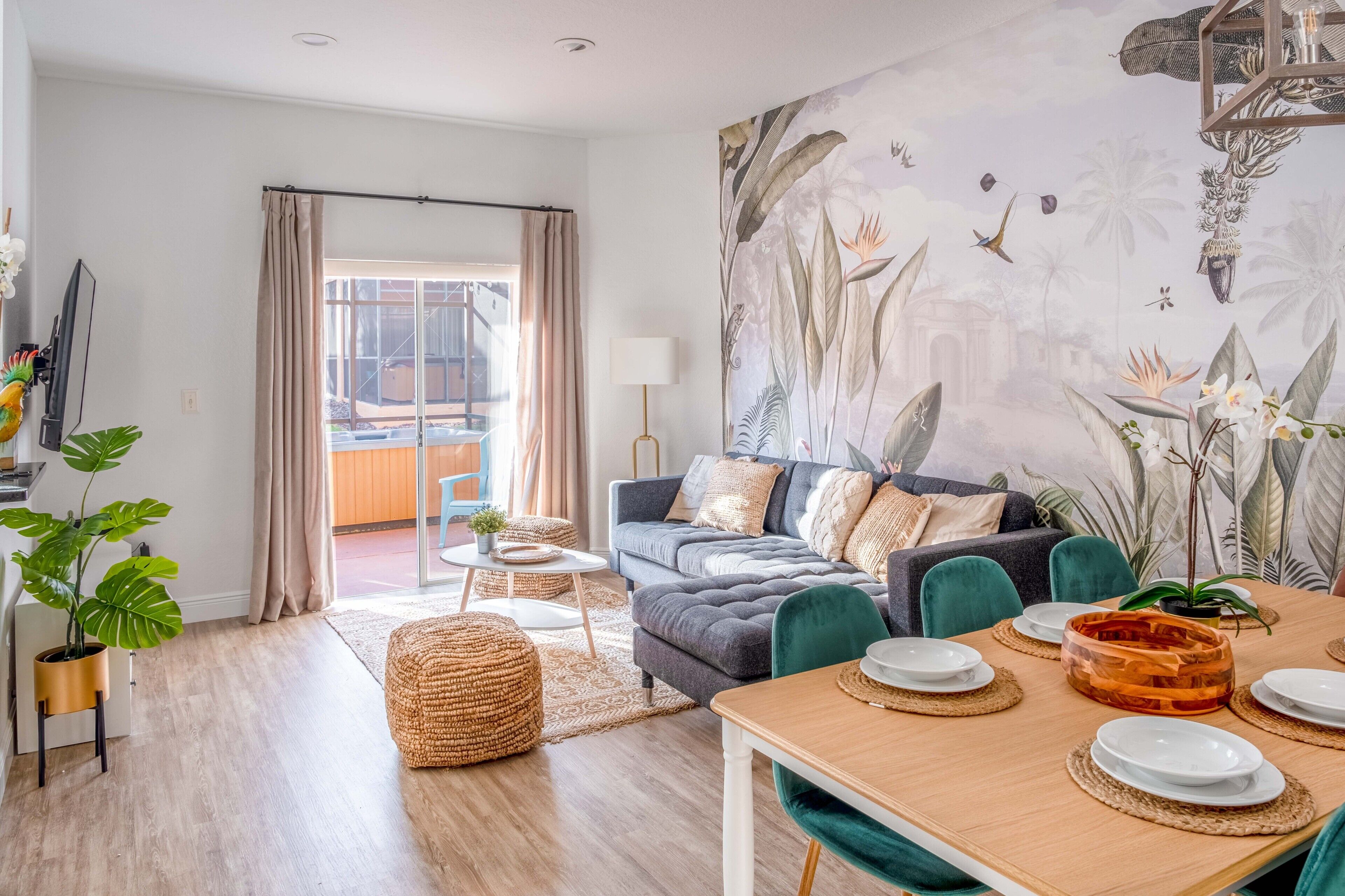 A modern living room with a grey sofa, teal chairs, a wooden dining table set for four, and a large tropical mural on the wall.