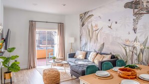 Ein modernes Wohnzimmer mit einem grauen Sofa, Türkis-farbenen Sesseln, einem Holzes Esstisch für vier Personen und einem großen tropischen Wandbild.