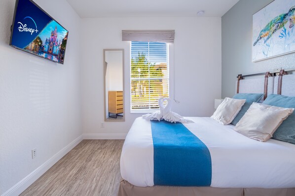 Une chambre d’hôtel avec un lit, une fenêtre avec des stores, un téléviseur Disney+ fixé au mur et un tableau.