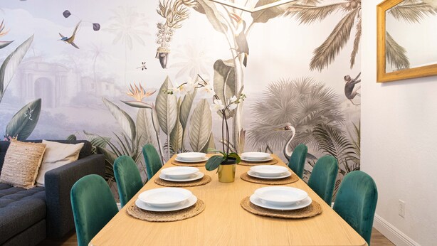 Un comedor con una mesa de madera dispuesta para seis personas, rodeada de sillas verdes, frente a una pared con un mural tropical.