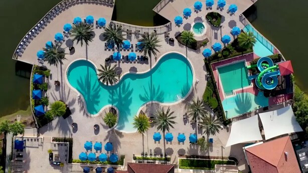 Eine Vogelperspektive zeigt ein Resort mit einem zentralen Swimmingpool, einer Wasserrutsche und mehreren Liegebereichen mit Sonnenschirmen.
