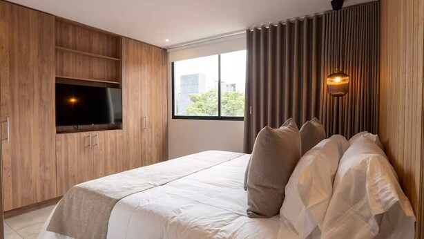 A modern bedroom with a large bed, a TV mounted on the wall, and a window with curtains. - Comfort and style in Medellin 301 (Medellín)