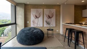 A modern kitchen with a large window, two wall-mounted flower paintings, a black bean bag chair, and a bar area with stools.