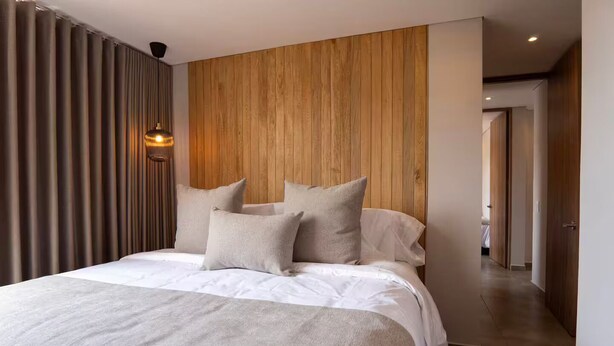 A modern bedroom with a wooden headboard, a large bed with white bedding, and a view of a hallway. - Perfect home in Medellín 501 (Medellín)