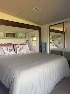 Une chambre avec un grand lit, un miroir, une fenêtre avec des stores et une bouche d’aération au plafond.