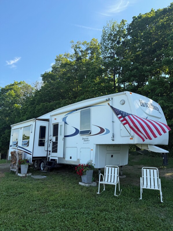 A white RV with 'Hitchhiker' branding and 'Discover America' text, parked on grass with a large American flag, surrounded by trees. - Pasture Perch: Renovated Camper, Farm Setting & Mountain Views Near Dream Park (Sharon Springs)