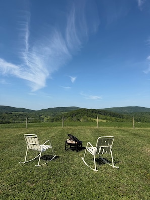Eine grüne Wiese mit zwei Schaukelstühlen und einer Feuerstelle, mit Blick auf eine hügelige Landschaft.