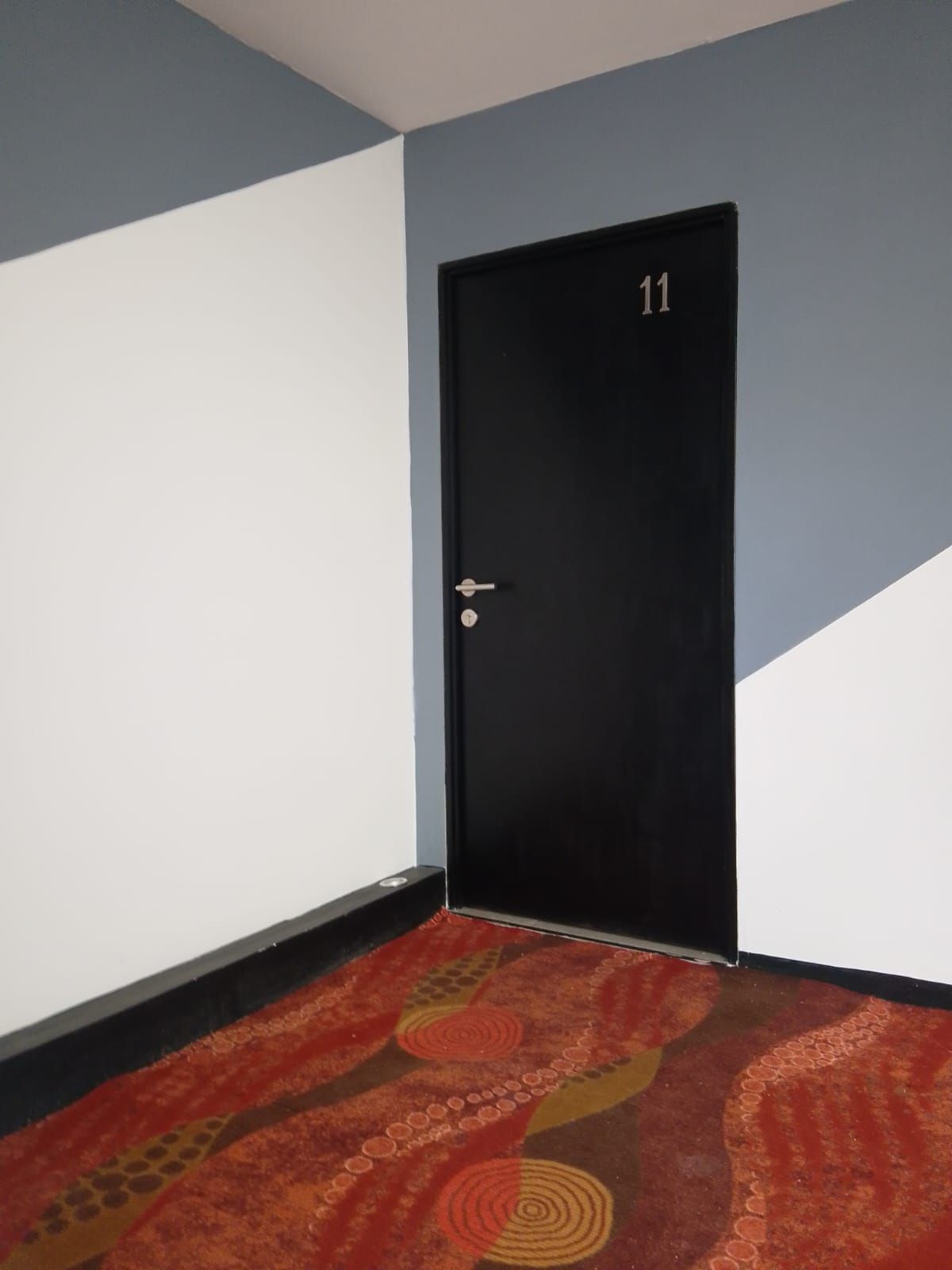 Una habitación con una puerta negra marcada con el número 11, paredes blancas y una alfombra roja con estampado.