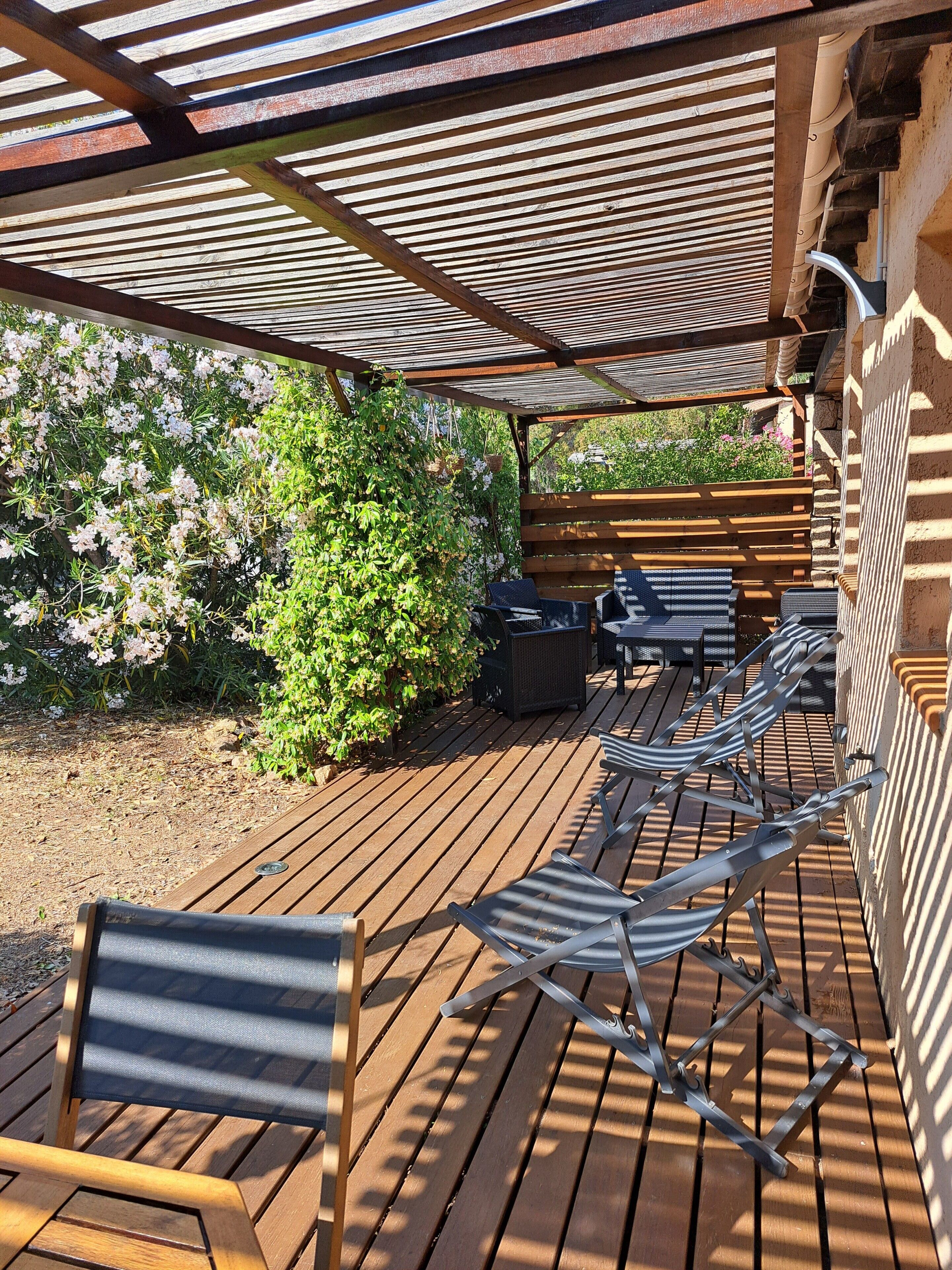 A wooden deck with metal chairs and a table, surrounded by greenery and a pergola.