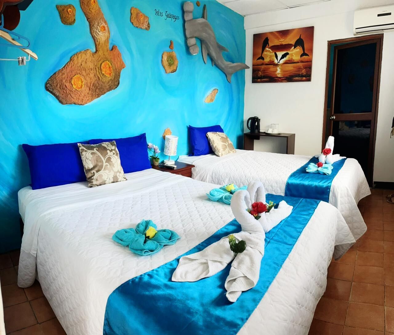 Une chambre d’hôtel avec deux lits, chacun recouvert d’une courtepointe bleue et blanche, et une fresque représentant un dauphin et des îles sur le mur.
