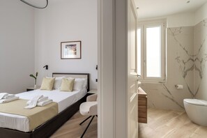 A modern bedroom with a bed, a bedside table, a chair, and a bathroom with a toilet and sink. - Domus Mea Suite (Florence)