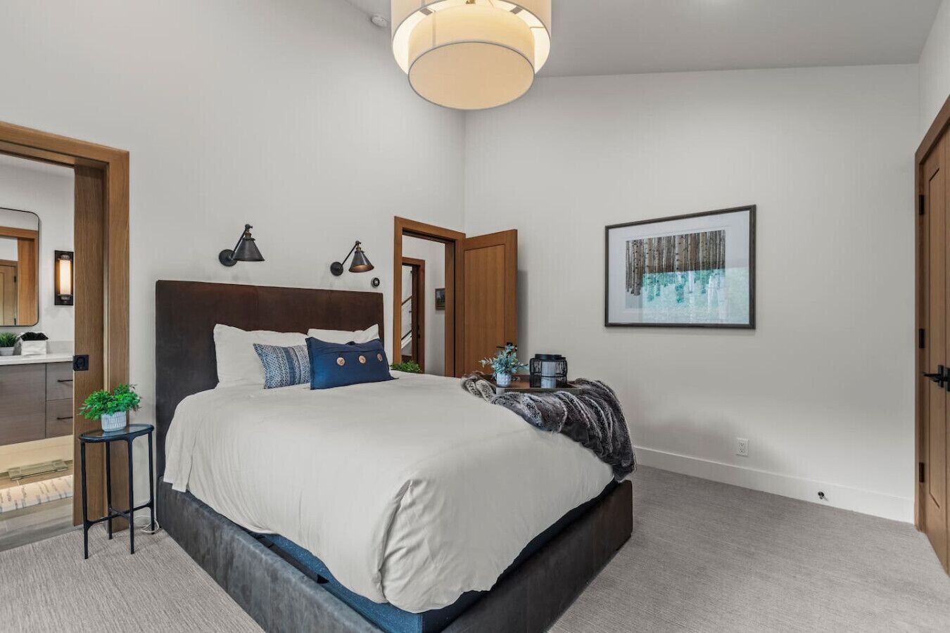 A modern bedroom with a large bed, bedside lamps, a small table with a plant, and a framed picture on the wall.