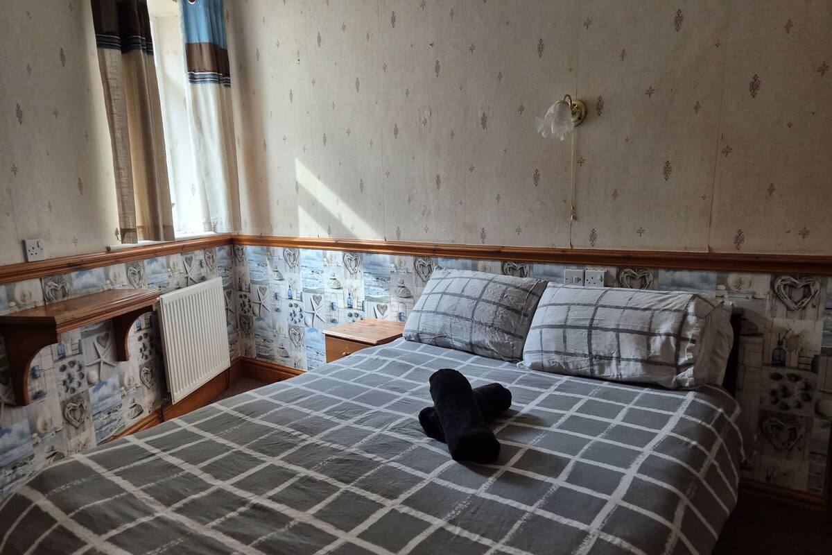 Un dormitorio con una colcha a cuadros, una mesita de noche, un radiador y un papel tapiz estampado.