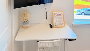 Una habitación con una televisión de pantalla plana montada en la pared, una mesita con un florero y un letrero de bienvenida, una silla blanca y una ventana con vista a una casa y montañas.