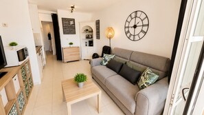 A living room with a sofa, a coffee table, a clock, and a kitchen area in the background. - Studio cabin center Holiday village Cap Estérel (Saint-Raphaël)