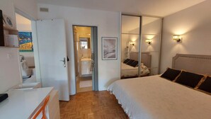 Une chambre à coucher avec un lit, des tables de chevet, un miroir et une salle de bain visible à travers une porte ouverte.