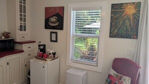 Uma cozinha compacta com micro-ondas, cafeteira e um pequeno frigorífico. Uma janela com persianas oferece luz natural, e um quadro é visível na parede.