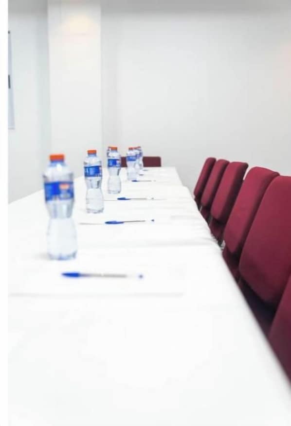 Un salón de conferencias con una larga mesa, sillas rojas y botellas de agua.