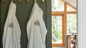 Two white robes hanging on a wooden rack against a green wall, leading to a room with a wooden deck and a view of greenery. - Modern+Hot Tub+Hocking Hills+ Near Waterfalls (Logan)