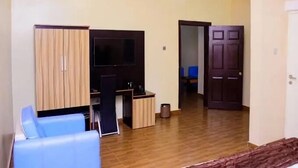 A room with a wooden floor, a TV mounted on the wall, a desk with a chair, and a wardrobe. - Rext  Apartment  (Abuja)