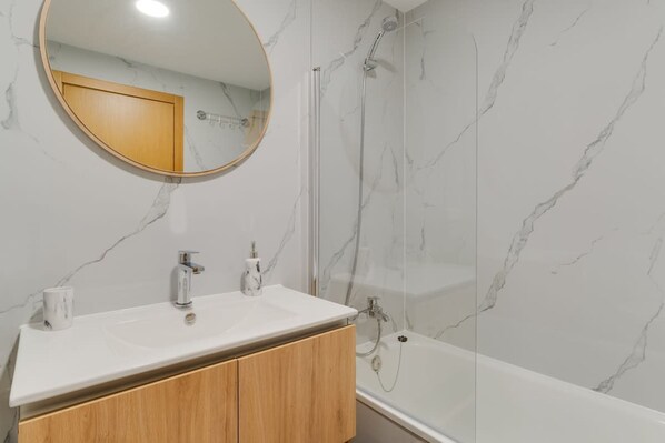 Un baño con lavabo de mármol blanco, armarios de madera, un espejo redondo y ducha con mampara de cristal.