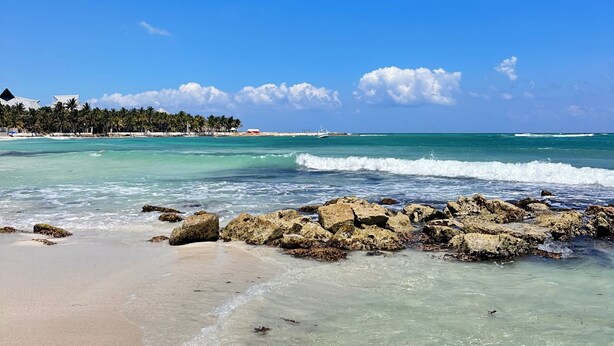 A beach with clear turquoise water, rocky shoreline, and a distant tree line.