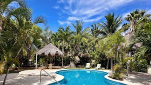 A swimming pool with a thatched-roof structure, surrounded by palm trees and lounge chairs.