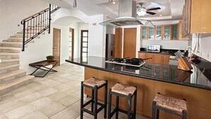 A spacious kitchen with a central island, bar stools, a staircase, and a ceiling fan.
