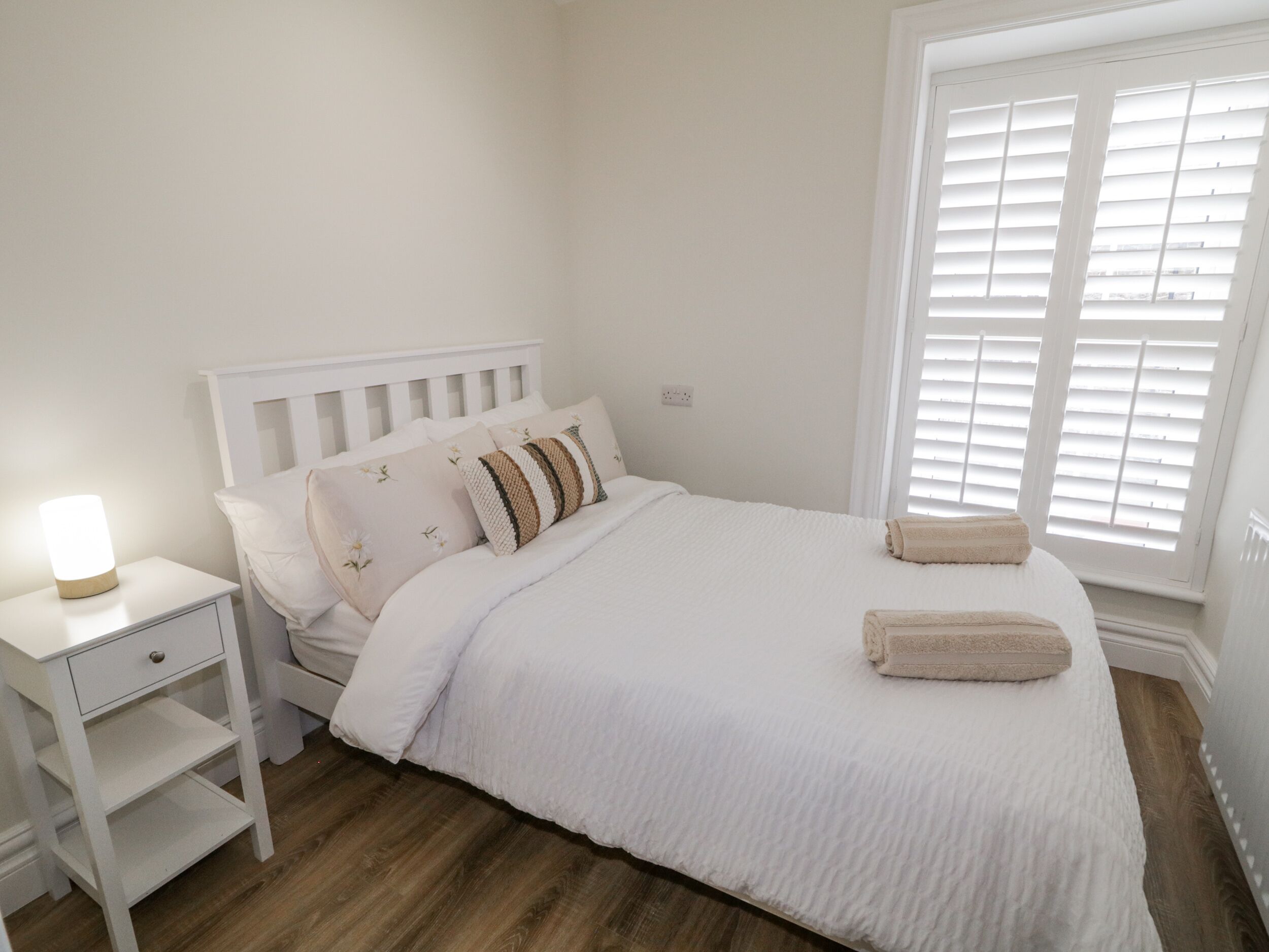 Una cama bien hecha con ropa blanca y un cabecero, una mesita de noche con una lámpara y una ventana con persianas.