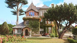 Una casa grande de dos pisos con fachada de piedra, una torre destacada y un jardín bien cuidado con diversas plantas y flores.