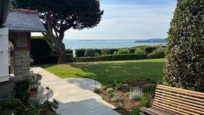 Un jardín ajardinado con un sendero de piedra que lleva a un banco, un árbol grande y vistas al mar.