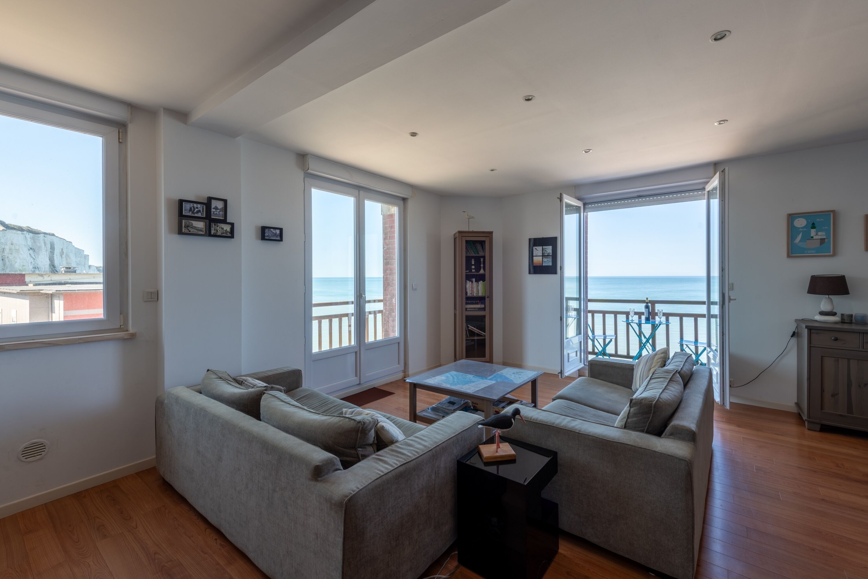 A living room with a sofa set, a coffee table, and a bookshelf. There are large windows with a view of the sea.
