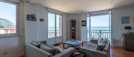 A living room with a sofa set, a coffee table, and a bookshelf. There are large windows with a view of the sea.