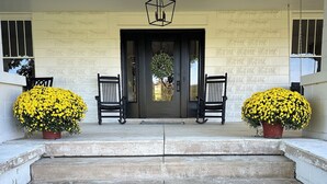 A set of concrete steps leading to a dark wooden door with a geometric light fixture above. There are two potted yellow flowers on either side of the door and two black rocking chairs facing each other. - Charming Farmhouse, Peaceful and Private (Hurricane Mills)
