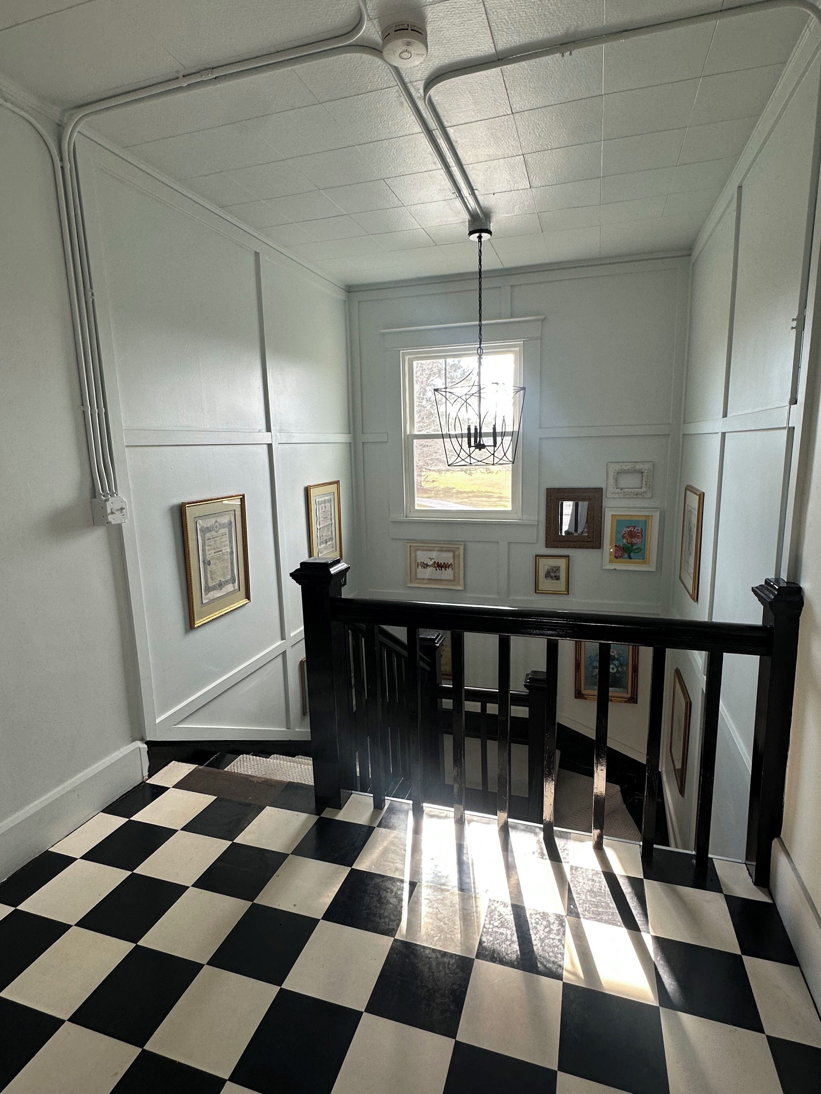 A hallway with a black and white checkered floor, a black railing, and framed pictures on the walls.