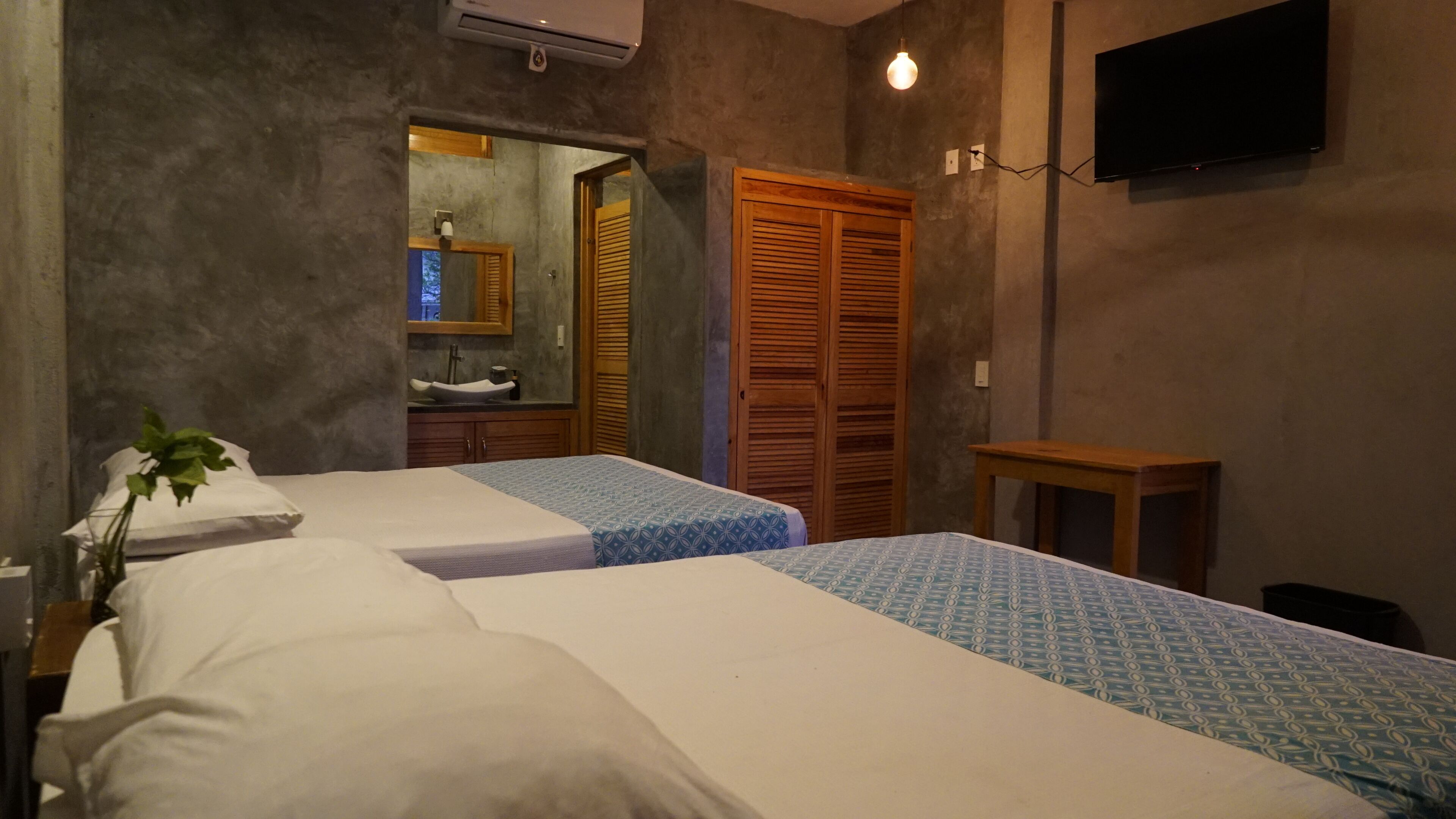 A bedroom with two beds, a wooden wardrobe, a small table, a TV, and a bathroom visible through an open door.