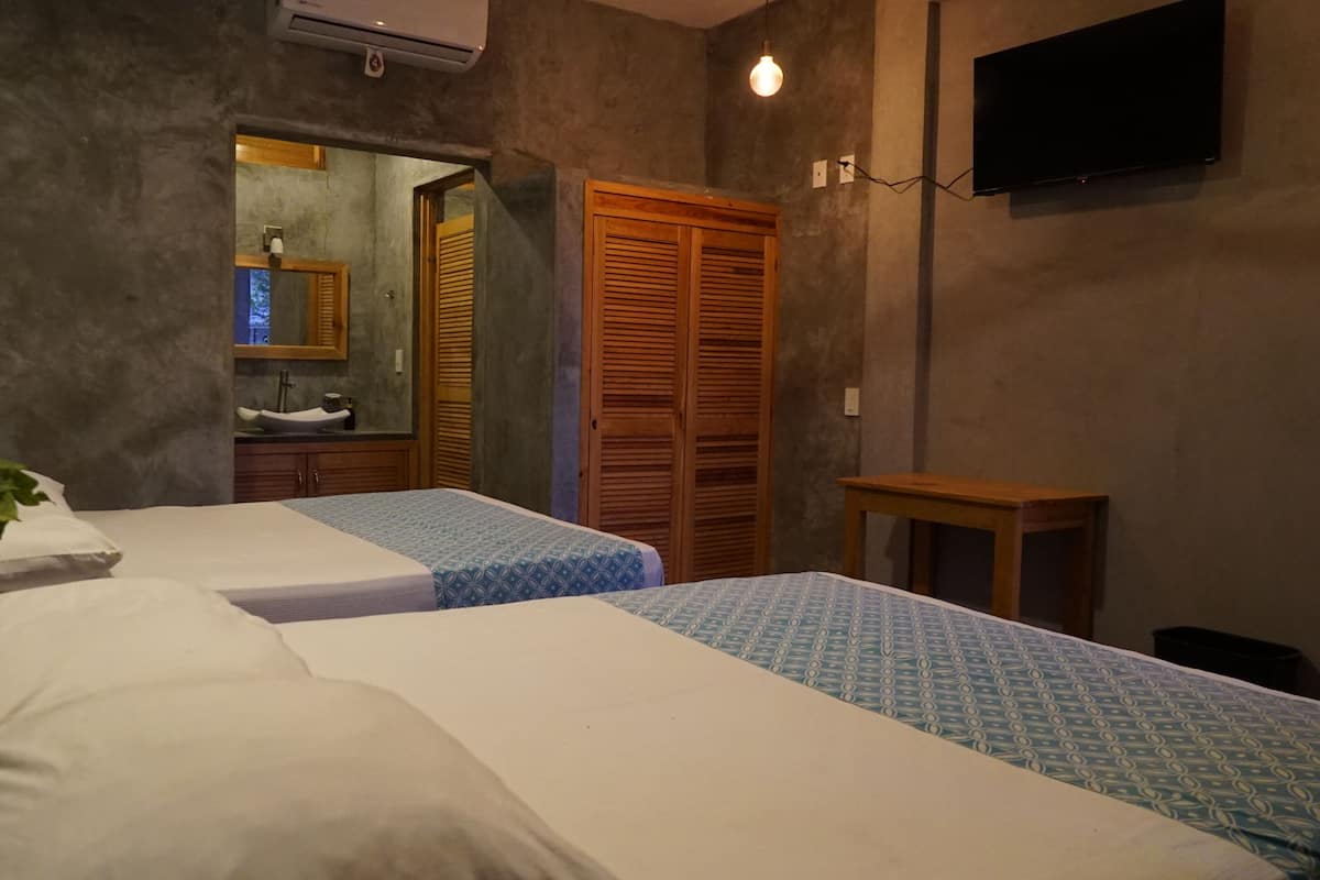Une chambre avec deux lits, une armoire en bois, une petite table, un téléviseur et une salle de bain visible à travers une porte ouverte.